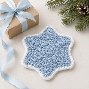 May include: A star-shaped crochet coaster in light blue yarn with a white border. The coaster features a textured, radiating stitch pattern. It is positioned near a brown gift box tied with a light blue satin ribbon and a pine branch with a pinecone.