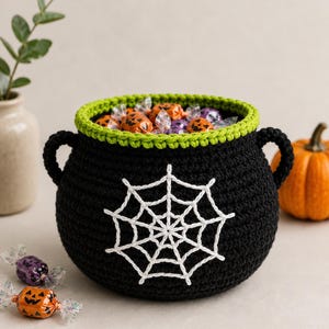 May include: A black crocheted cauldron basket with two side handles, featuring a bright green rim and a white embroidered spiderweb design on the front. The basket is filled with small orange and purple wrapped candies with pumpkin faces.
