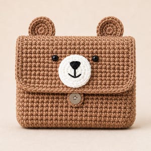 May include: A brown crocheted bear-shaped bag with a flap closure. The bag features a white and black bear face, two small ears, and a button closure. The handmade bag is a cute accessory.