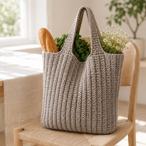 May include: A light beige, vertically ribbed crochet tote bag with two integrated handles. The bag is filled with a crusty baguette, a bunch of curly green kale, and a small bouquet of white daisies with yellow centres, creating a textured look.