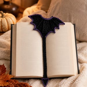 May include: A black crocheted bookmark featuring a bat wing design at the top and a pointed tail at the bottom, both edged with a vibrant purple yarn. The bookmark is placed vertically inside an open book with blank white pages.