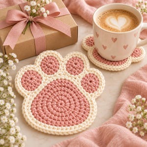 Pode incluir: Dois porta-copos de crochê feitos à mão em formato de pata de animal, com centro e almofadas em rosa antigo contornados com fio cor de creme. Um porta-copos está plano, enquanto o outro sustenta uma caneca de cerâmica branca com corações.