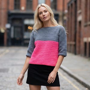 Könnte beinhalten: Ein grau-rosa Häkeltop mit Dreiviertelärmeln. Das Oberteil ist horizontal geteilt, wobei der obere Teil grau und der untere Teil leuchtend rosa ist. Der Häkelstich erzeugt ein strukturiertes Aussehen.