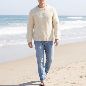 May include: A person wearing a cream-colored, crew-neck sweater and light blue jeans walks on a sandy beach. The sweater has a textured knit, and the jeans are a classic, straight-leg style. The background shows the ocean and a clear sky.