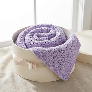 May include: A rolled-up, light purple crocheted blanket rests inside a round, cream-colored gift box with a matching ribbon. The blanket's open weave pattern is visible, and a portion of the blanket extends over the edge of the box.