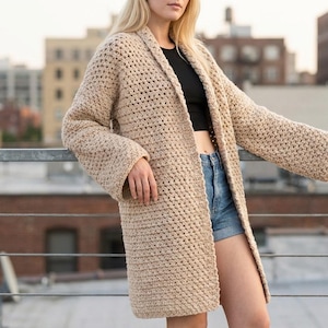 May include: A long, beige crochet cardigan with a shawl collar and long sleeves. The open-front design features a textured stitch pattern. The cardigan is knee-length and appears to be made of a soft, chunky yarn.