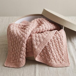 Op de afbeelding: Een zachtroze gebreide deken met een textuurpatroon, gedeeltelijk opgevouwen in een ronde, lichtgekleurde doos. De deken heeft een diamantachtig ontwerp op een hoek. Ideaal voor een baby.