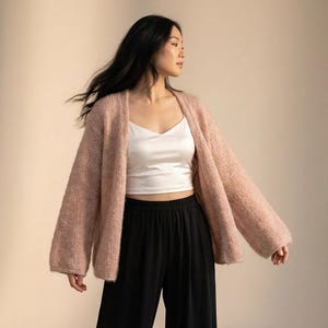 May include: A woman wearing a light pink, open-front cardigan with long sleeves. Underneath, she wears a white camisole and black pants. The cardigan is a soft, textured knit, and the outfit is set against a neutral background.