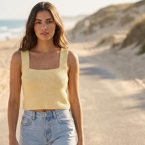 May include: A light yellow, square-neck crochet top with scalloped edges. The top is paired with light blue denim jeans. The background shows a beach scene with a road, sand, and the ocean.