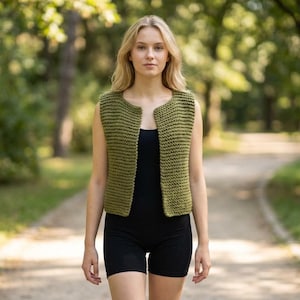 May include: Olive green knitted vest with a round neckline and sleeveless design. The open-front vest is worn over a black top and paired with black shorts. The image is taken outdoors with a blurred green background.