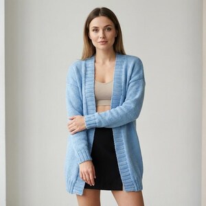 May include: A light blue, open-front cardigan sweater. The long-sleeved sweater is knee-length and has a ribbed texture. The person in the image is wearing a beige top and a black skirt. The cardigan is a soft, comfortable knit.