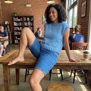 May include: A person wearing a light blue knit top and denim shorts sits on a wooden table. The person is in a cafe with a brick wall and bookshelves. A coffee cup and book are on the table.