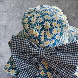 May include: A sun hat with a blue and white daisy print and a large gingham bow. The hat has a wide brim and a floral pattern with white daisies and yellow centres. The bow is a blue and white checkered pattern.