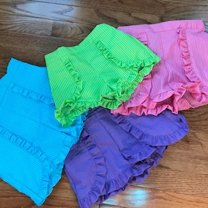 May include: Four pairs of ruffled shorts in various colors: light blue, green, pink, and purple. The shorts have a seersucker stripe pattern and a ruffled hem. The shorts are arranged on a wooden surface.