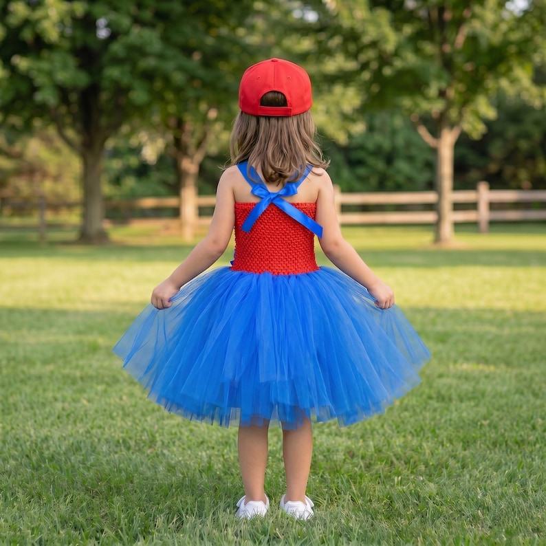 Op de afbeelding: Een rode honkbalpet gedragen bij een jurk met een rood gehaakt lijfje en een volle blauwe tule rok. Het lijfje heeft blauwe lintbandjes die op de rug kruisen en in een strik worden vastgemaakt. De jurk wordt gedragen met witte schoenen.