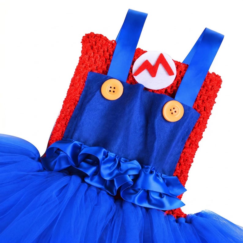 Op de afbeelding: Een rode gehaakte jurk met een blauwe satijnen tuinbroek-overlay, twee gele knopen, blauwe satijnen schouderbanden, een witte ronde patch met een rode letter M en een gelaagde blauwe tule rok met een geplooide satijnen rand.