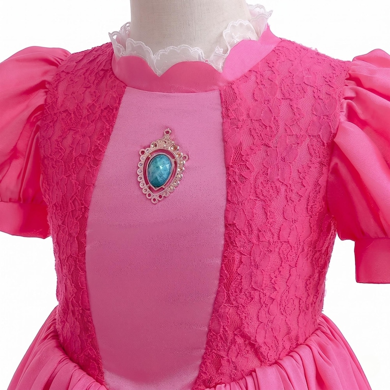 May include: A pink princess dress with a jewel-like brooch. The bodice features a lace overlay and a satin panel. The dress has short, puffy sleeves and a white lace collar. The skirt is a flowing, pleated design.