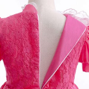 May include: A close-up of a pink dress with a white lace collar and a zipper down the back. The dress features a floral lace overlay on the bodice and sleeves. The dress is made of a soft, flowing fabric.