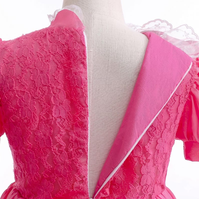 May include: A close-up of a pink dress with a white lace collar and a zipper down the back. The dress features a floral lace overlay on the bodice and sleeves. The dress is made of a soft, flowing fabric.