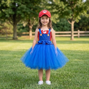 Op de afbeelding: Een rode honkbalpet met een wit M-logo en een blauw-rode tutu-jurk met een rood gehaakt lijfje, blauwe bandjes met gele knopen en een volle blauwe tule rok. Het lijfje is voorzien van een witte ronde patch met een rood M-embleem.