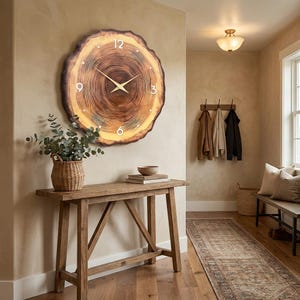 May include: A large circular wall clock featuring a natural wood grain pattern that mimics a tree cross-section. The clock face displays white numerals at the 12, 3, 6, and 9 o'clock positions, with thin gold-toned hands centered on the wood surface.