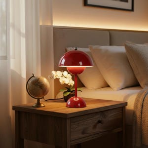 May include: A glossy red mushroom-shaped table lamp with a rounded dome shade and a slender stem base. The lamp features a smooth, reflective finish and a circular base, providing a focused downward light source on a wooden bedside table.