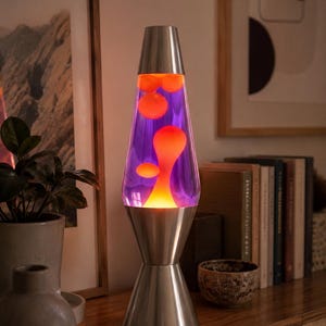 May include: A retro lava lamp with a silver base and top, and a clear glass bottle filled with purple liquid and orange wax blobs. The lamp is on a wooden surface, with books and decor in the background.