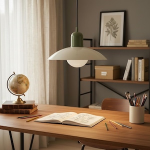 May include: A stylish interior with a green and white pendant lamp suspended over a wooden desk. A globe, books, and art supplies are arranged on the desk, with a framed botanical print on a shelf in the background. The room is illuminated by natural light.