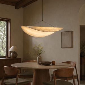Puede incluir: Una lámpara colgante grande, curva y en forma de media luna con una pantalla texturizada, de color blanco roto y translúcida. La luz está suspendida por cables negros finos del techo, emitiendo un brillo suave y cálido hacia abajo.