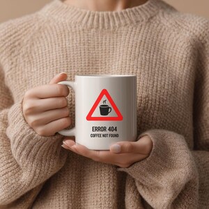 May include: A white ceramic mug featuring a red triangular warning sign icon with a black coffee cup graphic inside. Below the icon, the text reads "ERROR 404" and "COFFEE NOT FOUND" in black sans-serif lettering. The mug has a curved handle.