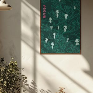 May include: A framed poster featuring a dark green illustration of a tree with numerous white spirit-like figures. The Japanese text "もののけ姫" is in pink on the left side. The artwork is displayed on a white wall.