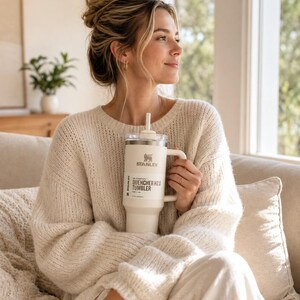 May include: A cream-coloured Stanley Quencher H2.0 Tumbler with a handle and straw. The tumbler has the Stanley logo and product information printed on the side. The person is wearing a cream-coloured jumper and sitting on a sofa.