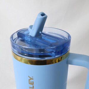 Op de afbeelding: Een lichtblauwe ge&iuml;soleerde beker met een handvat en een doorzichtig, blauw getint deksel. Het deksel heeft een flip-top tuit en een goudkleurige band aan de bovenkant. Op de beker staat het woord "LEY" gedrukt.