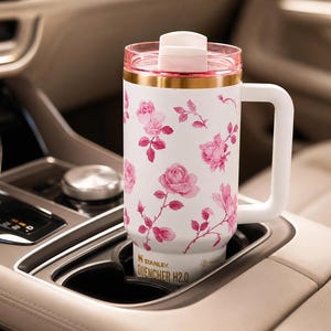 May include: White Stanley Quencher H2.0 travel mug with a pink floral design. The mug has a white handle, a pink lid, and a gold band. The mug is in a car's cup holder.