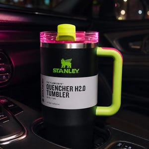 Pu&ograve; includere: Bicchiere Stanley Quencher H2.0 nero e verde neon con manico, in un portabicchieri per auto. Il bicchiere ha un manico e un coperchio verde neon, con il logo Stanley e "QUENCHER H2.0 TUMBLER" stampato. Contiene 1,18 L.