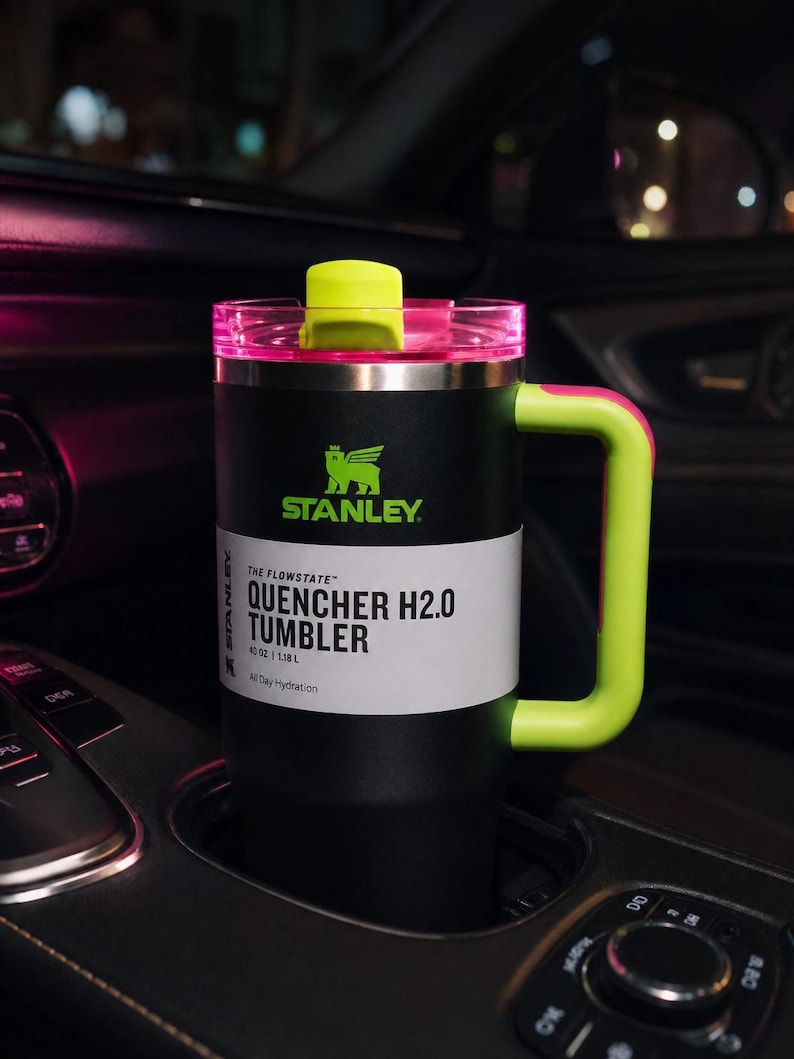 Pu&ograve; includere: Bicchiere Stanley Quencher H2.0 nero e verde neon con manico, in un portabicchieri per auto. Il bicchiere ha un manico e un coperchio verde neon, con il logo Stanley e "QUENCHER H2.0 TUMBLER" stampato. Contiene 1,18 L.