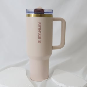 May include: A light beige Stanley travel mug with a handle. The mug has a clear lid with a dark red center and gold trim. The Stanley logo is printed vertically on the side of the mug.