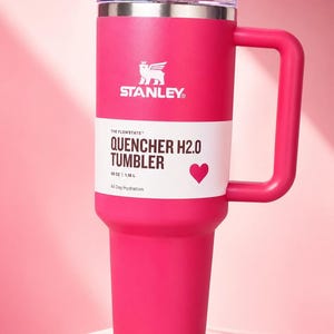 Puede incluir: Un vaso Stanley Quencher H2.0 de color rosa brillante con asa. El vaso tiene una tapa transparente y una banda blanca con el texto "QUENCHER H2.0 TUMBLER" y un s&iacute;mbolo de coraz&oacute;n. Contiene 1,18 L y est&aacute; dise&ntilde;ado para la hidrataci&oacute;n durante todo el d&iacute;a.