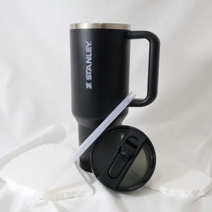 May include: Black Stanley travel mug with a handle, lid, and straw. The mug has a silver rim and the Stanley logo in white. The lid is black with a sliding closure. The straw is clear.