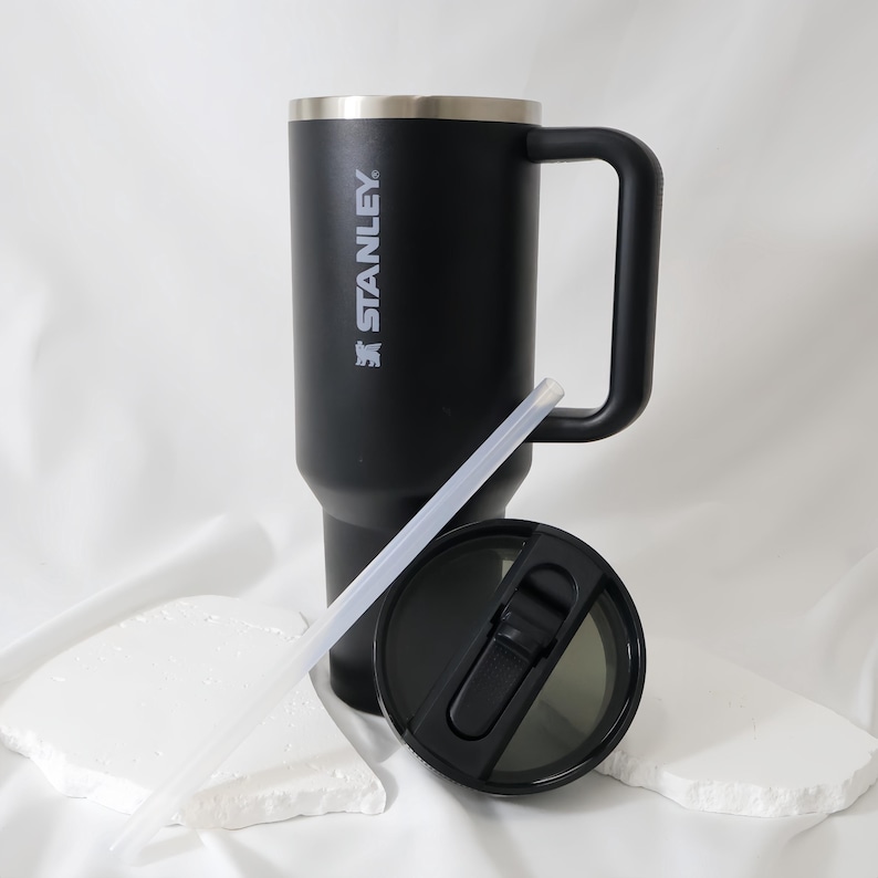 May include: Black Stanley travel mug with a handle, lid, and straw. The mug has a silver rim and the Stanley logo in white. The lid is black with a sliding closure. The straw is clear.