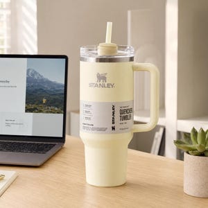 May include: A cream-colored Stanley Quencher tumbler with a handle and straw sits on a wooden desk. The tumbler has the Stanley logo and product information labels. A laptop displaying a website and a small potted succulent are also on the desk.