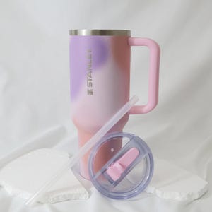Op de afbeelding: Een Stanley-beker met een handvat, rietje en deksel. De beker heeft een gradi&euml;ntontwerp van roze, paars en wit. Het handvat en de dekselaccenten zijn roze. De beker is ontworpen om een grote hoeveelheid vloeistof te bevatten.