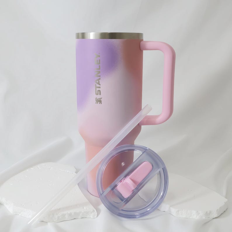 Op de afbeelding: Een Stanley-beker met een handvat, rietje en deksel. De beker heeft een gradi&euml;ntontwerp van roze, paars en wit. Het handvat en de dekselaccenten zijn roze. De beker is ontworpen om een grote hoeveelheid vloeistof te bevatten.