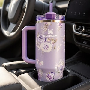 May include: A lavender Stanley tumbler with a floral design, featuring a handle and a straw. The tumbler is in a car's cup holder. The Stanley logo is visible on the front.