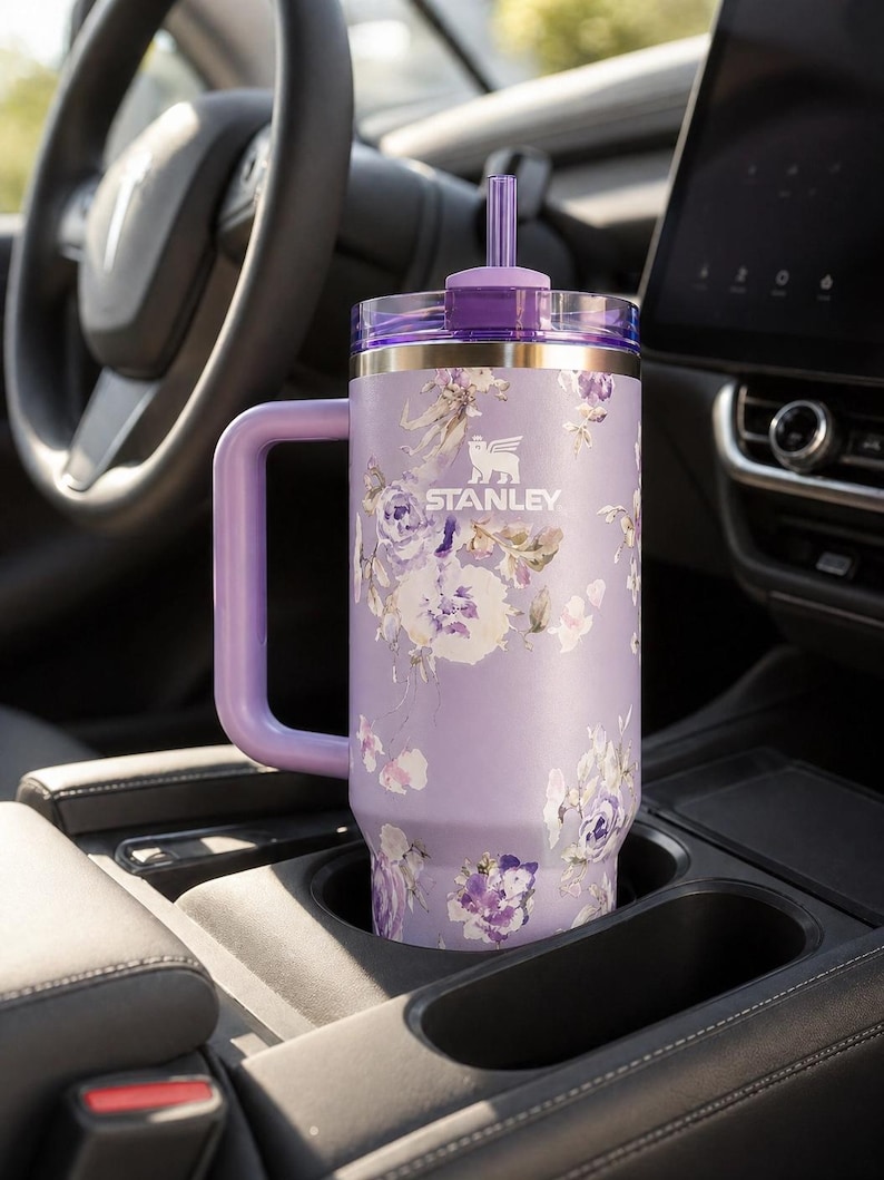 May include: A lavender Stanley tumbler with a floral design, featuring a handle and a straw. The tumbler is in a car's cup holder. The Stanley logo is visible on the front.