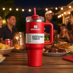 K&ouml;nnte beinhalten: Ein roter Stanley Quencher H2.0 Becher mit Griff und Strohhalm, auf einem Holztisch. Der Becher hat ein wei&szlig;es Etikett mit schwarzem Text. Im Hintergrund essen Leute im Freien.