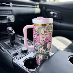 Może przedstawiać: R&oacute;żowy i biały kubek Stanley Loveshackfancy z uchwytem, ​​z powtarzającym się wzorem postaci z kresk&oacute;wek. Kubek znajduje się w uchwycie na kubek w samochodzie. Ma złotą obw&oacute;dkę i logo Stanley.