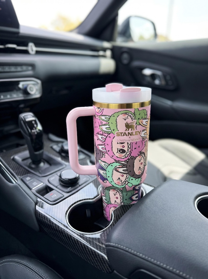 Może przedstawiać: R&oacute;żowy i biały kubek Stanley Loveshackfancy z uchwytem, ​​z powtarzającym się wzorem postaci z kresk&oacute;wek. Kubek znajduje się w uchwycie na kubek w samochodzie. Ma złotą obw&oacute;dkę i logo Stanley.