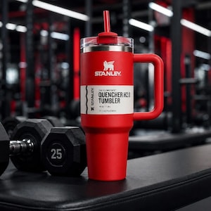 K&ouml;nnte beinhalten: Ein roter Stanley Quencher H2.0 Tumbler mit Griff und Strohhalm steht auf einer schwarzen Oberfl&auml;che neben Hanteln. Der Becher hat ein wei&szlig;es Etikett mit schwarzem Text und dem Stanley-Logo. Der Hintergrund ist ein Fitnessstudio.