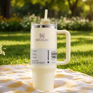 May include: Cream-colored Stanley Quencher Tumbler with a handle and straw, set on a yellow and white striped picnic blanket. The tumbler has the Stanley logo and product information on a white label. The background features a grassy area and trees.